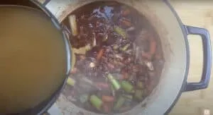 Pouring broth into a pot of simmering vegetables for a hearty stew recipe.